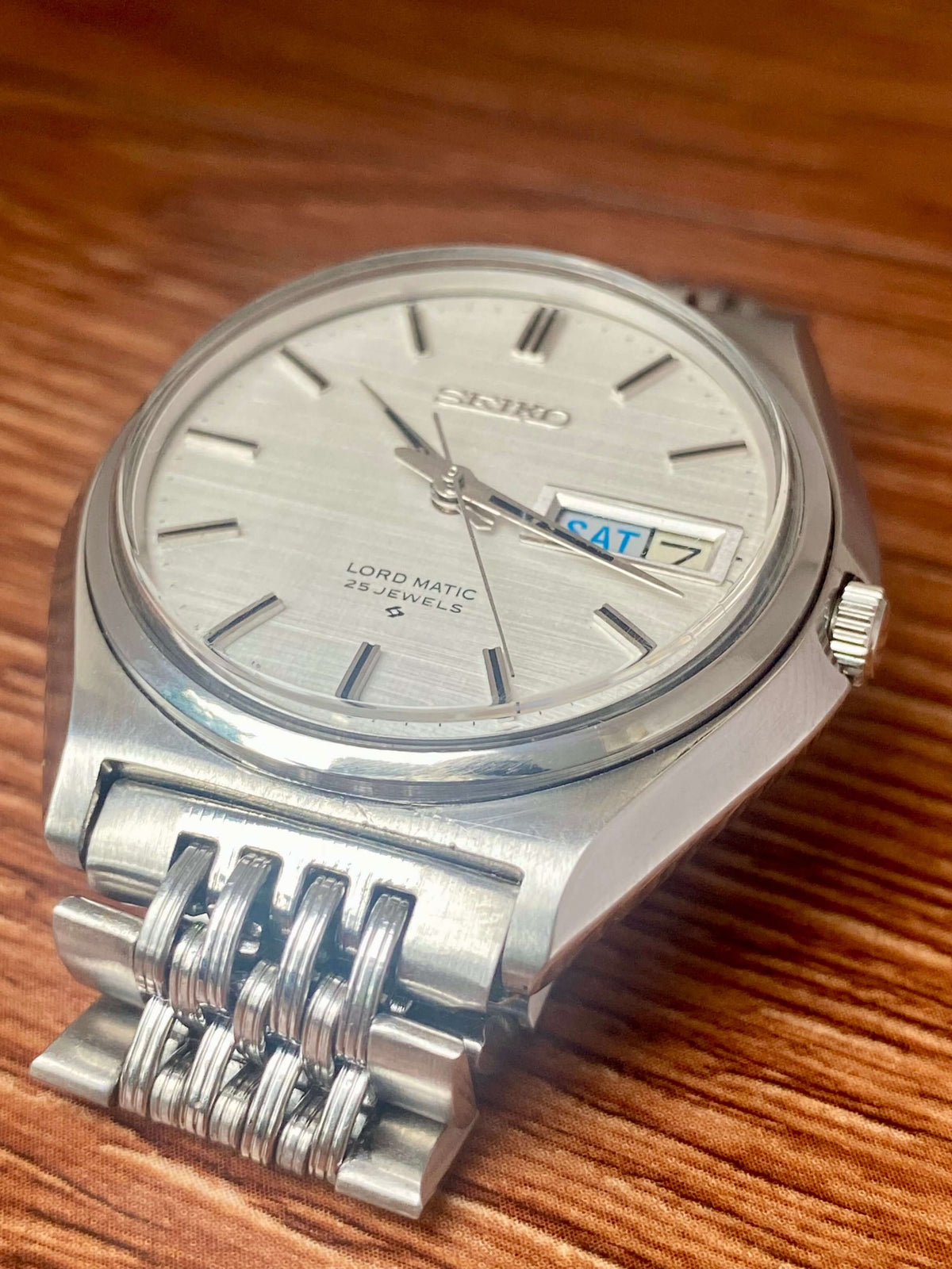 Vintage Seiko Lord Matic 5606-7010 on wooden surface, showcasing original white linen dial and unique bracelet.