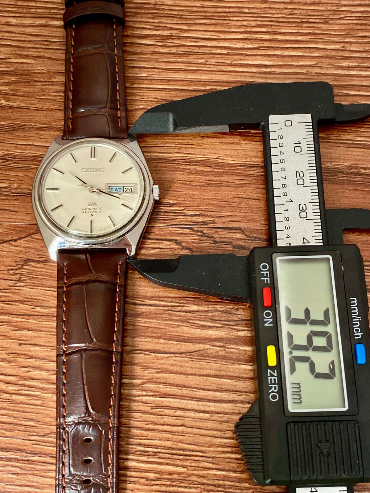 Vintage Seiko Lord Matic 5606-7000 watch displayed with caliper measuring size on a wooden surface.